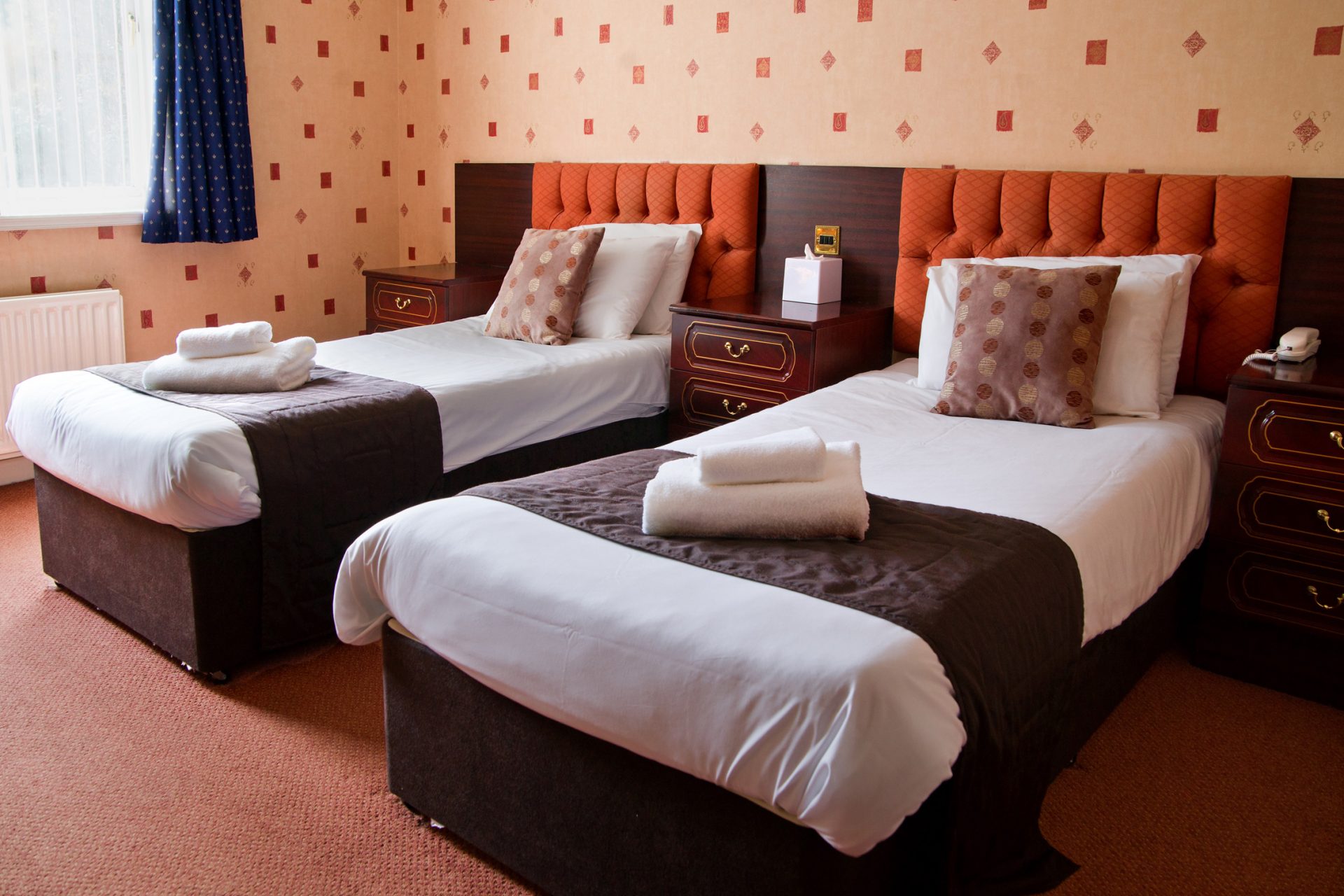 A neatly made hotel beds with a tufted orange headboards and patterned cushions.
