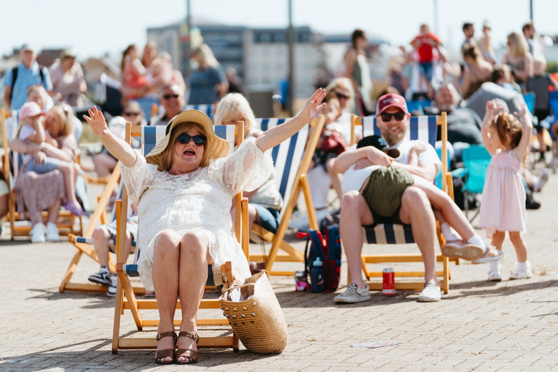 A woman in a sunhat sits on a deck chair with arms raised and mouth open, surrounded by people relaxing on chairs outdoors on a sunny day. A man and a young girl are nearby, and a crowd is in the background.