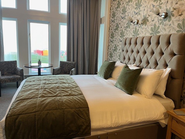 A cozy hotel room with a large bed, green bedding and pillows, patterned wallpaper, two chairs by tall windows, and a small round table overlooking a colourful view outside.