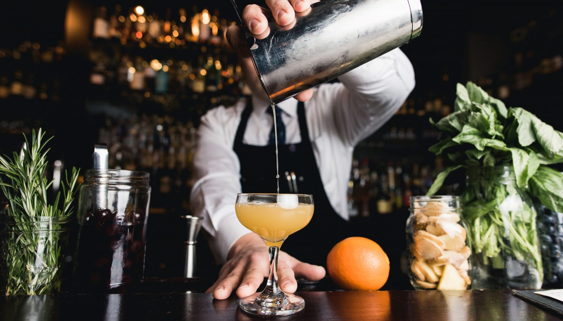 Man wearing shirt, tie and apron pours a cocktail from a metal shaker.
