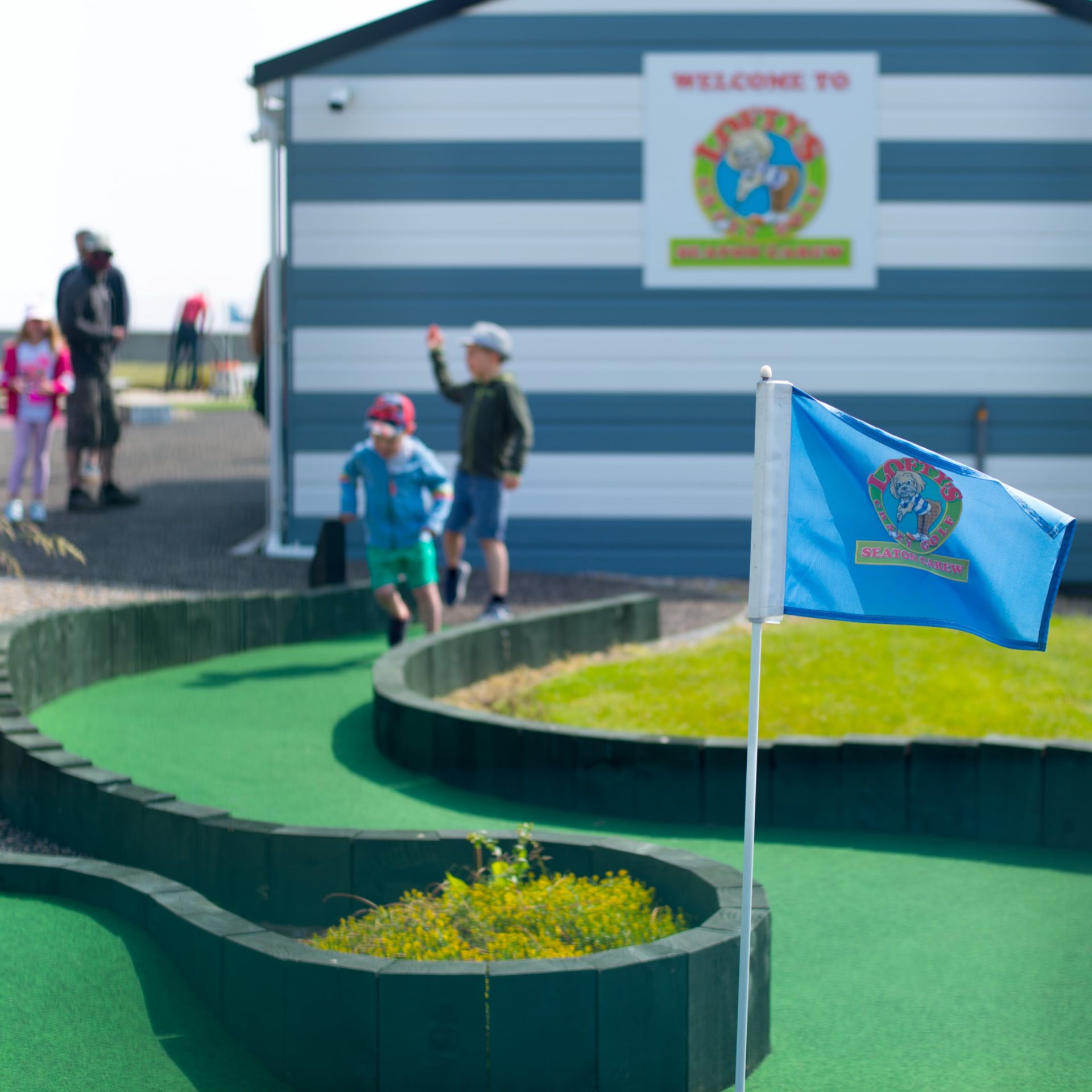A blue flag marks a mini golf course with children playing in the background near a striped building that has a colourful welcome sign.