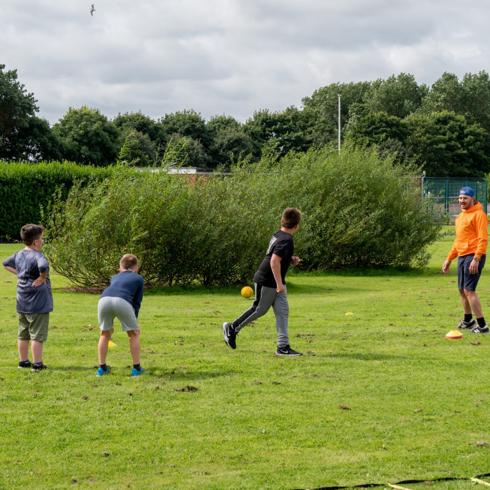 Four boys play a ball game on a grassy field, with trees and bushes in the background. One boy, dressed in an orange shirt and cap, appears to be instructing or coaching the others. It is a cloudy day.