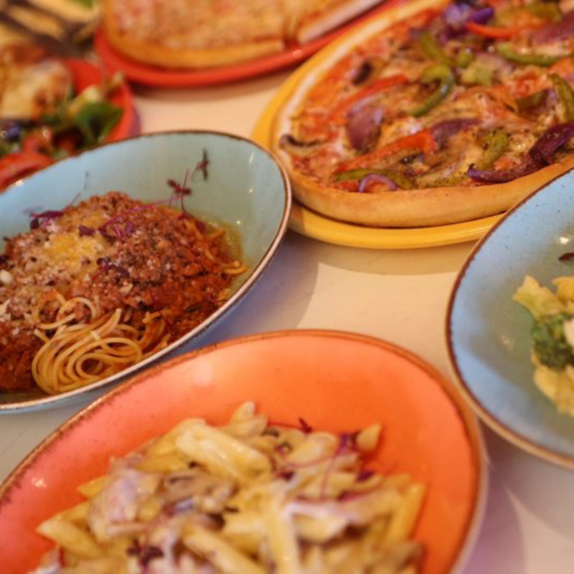 Various Italian dishes, including pizzas, spaghetti bolognese and creamy penne on colourful plates.