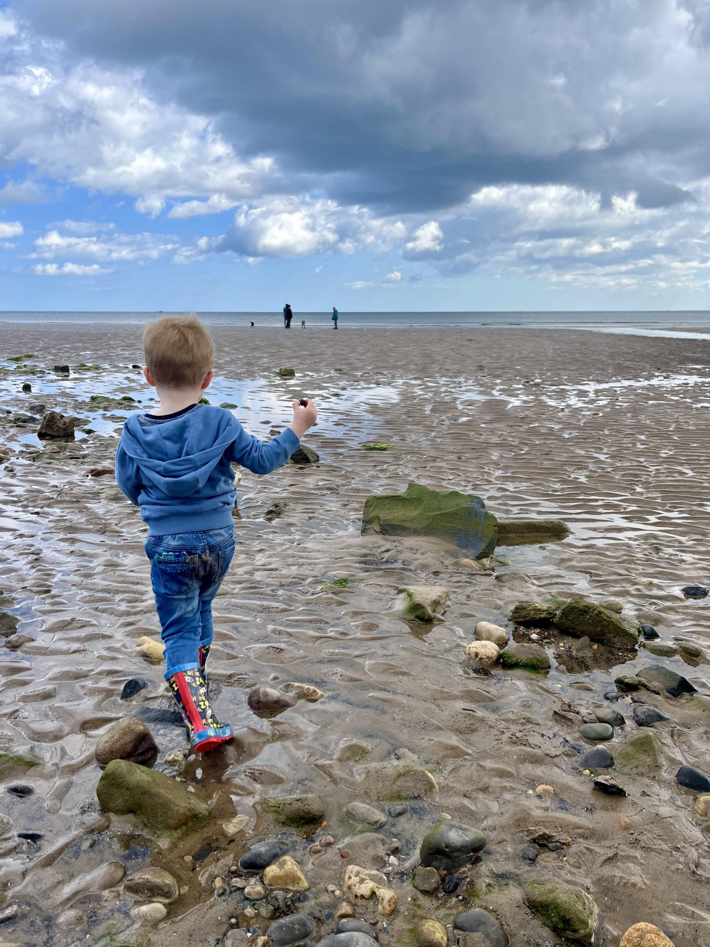 A young child wearing a blue hoodie and colourful boots walks on a rocky, sandy beach toward the sea under a cloudy sky. There are a few people in the distance near the water.