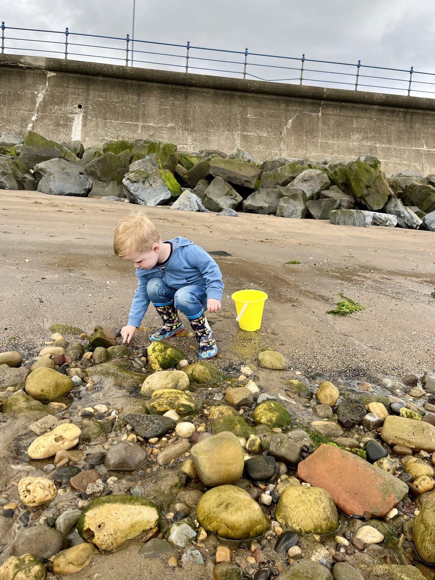 A young child in a blue hoodie and patterned rain boots crouches on a rocky beach, picking up stones near a yellow bucket. Large rocks and a concrete sea wall are visible in the background under a cloudy sky.