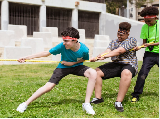Three boys outdoors are playing tug of war, pulling a yellow rope. They are wearing casual clothes and headbands, and are leaning back with effort on a grassy field. Concrete steps are visible in the background.