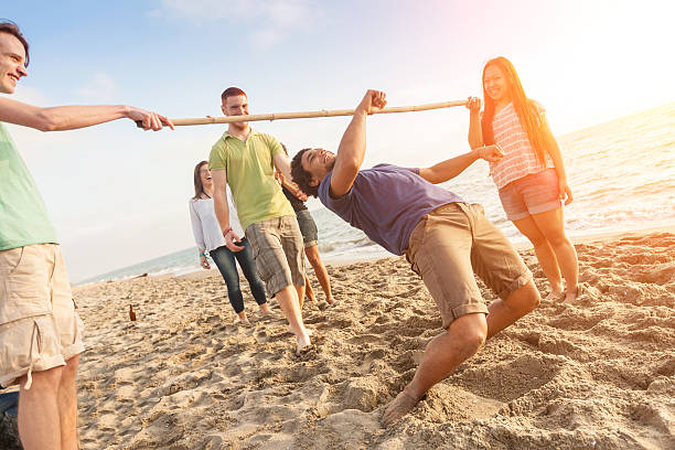 A group of young adults play limbo on a sandy beach near the ocean, with one person bending backward under a pole while others watch and smile in the sunlight.