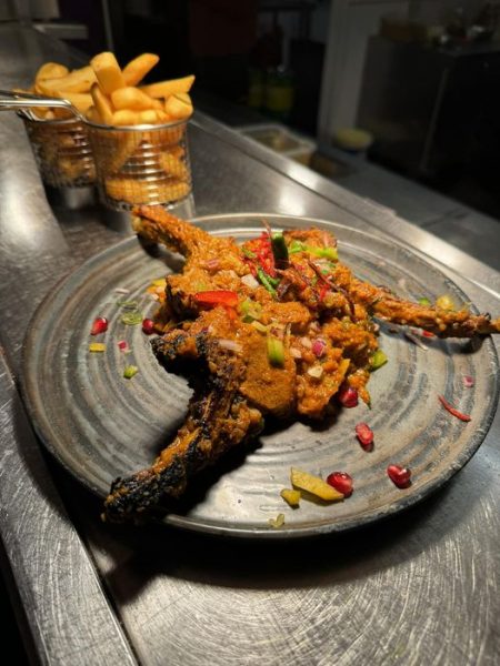 Grilled lamb chops topped with colourful vegetables and pomegranate seeds, served on a dark plate. In the background, a metal basket holds thick-cut fries on a restaurant counter.