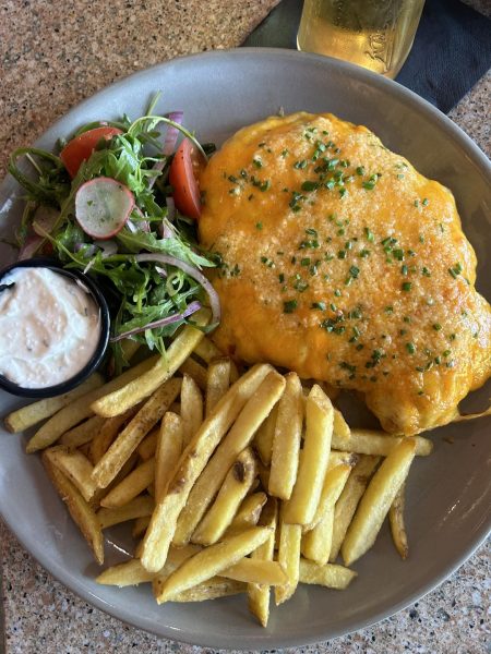 A plate with crispy fries, a fresh salad with radish and tomatoes, a cheddar cheese-covered parmo garnished with chives, and a small dish of creamy dipping sauce.