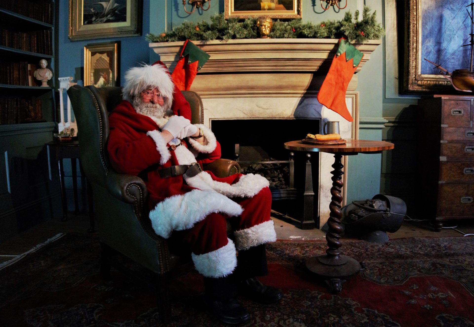 Santa Claus sits in an armchair by a fireplace in a cozy, decorated room. Two filled Christmas stockings hang on the mantel, and a plate of cookies and a glass of milk are on a nearby table.