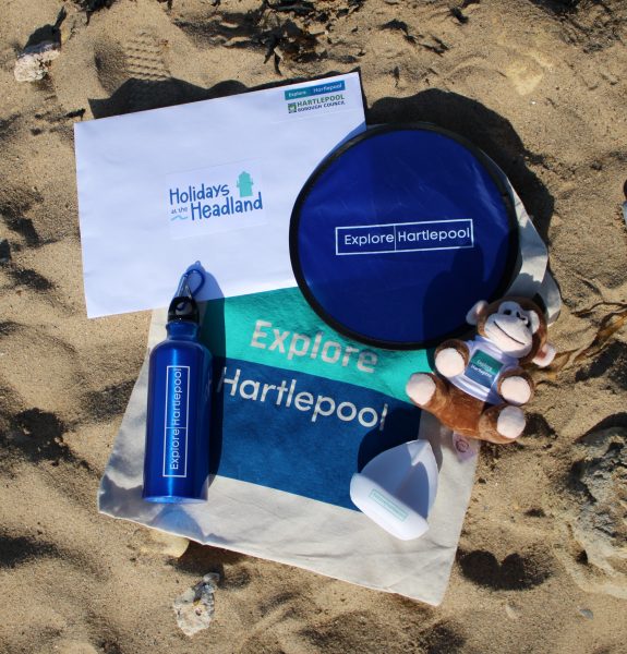 Promotional Explore Hartlepool items on sand, including a tote bag, water bottle, frisbee, small plush monkey, mini sunscreen, and a Holidays at the Headland envelope.