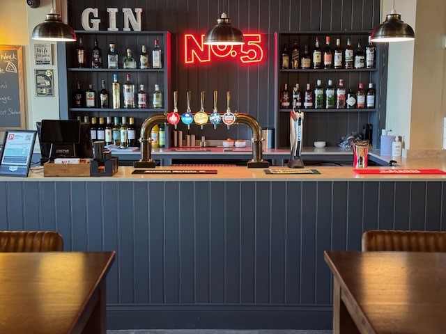 A bar with a dark wood counter and shelving stocked with bottles of alcohol. A neon sign reads No.5 above the bar, with GIN letters on the upper left shelf. Beer taps and glassware are on the counter.