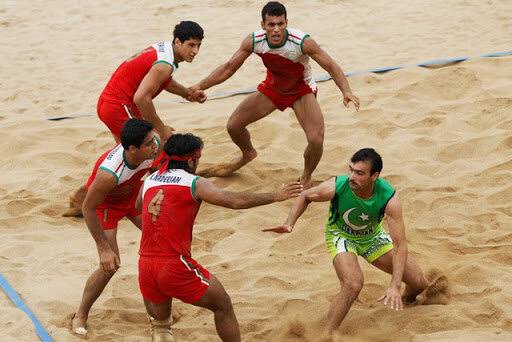 Five men play kabaddi on a sandy court. Four players in red uniforms surround a player in a green uniform, who appears to be evading their attempts to tag or tackle him.