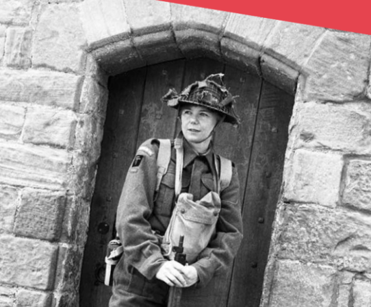 A person dressed in historical military uniform stands in front of a stone doorway, holding a rifle.