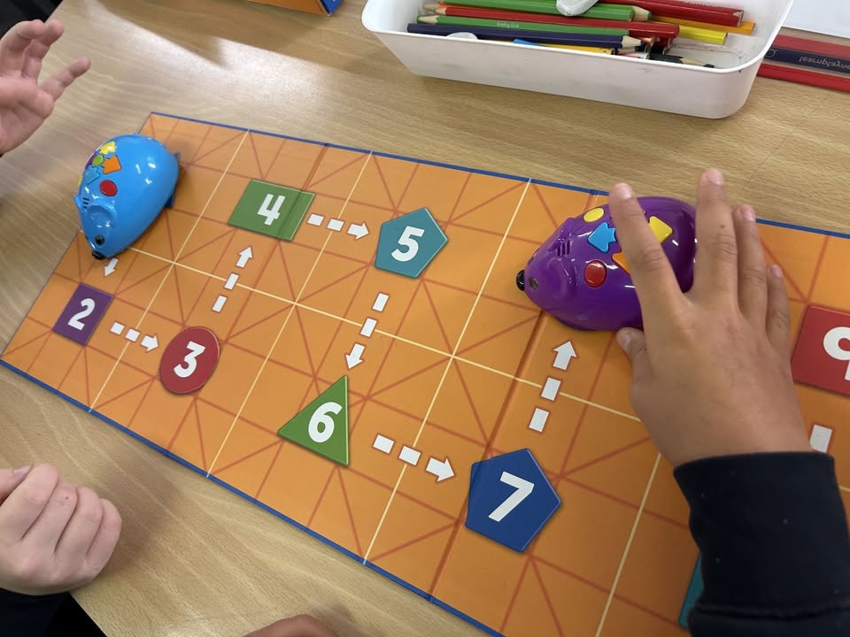 Two children are playing with blue and purple toy mice on a colorful board game with numbered spaces, arrows, and shapes. A tray of colored pencils is visible at the top of the table.