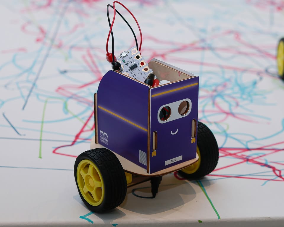A small, purple, wheeled robot with a smiling face and exposed electronic components sits on a white surface covered in colorful scribbled lines.