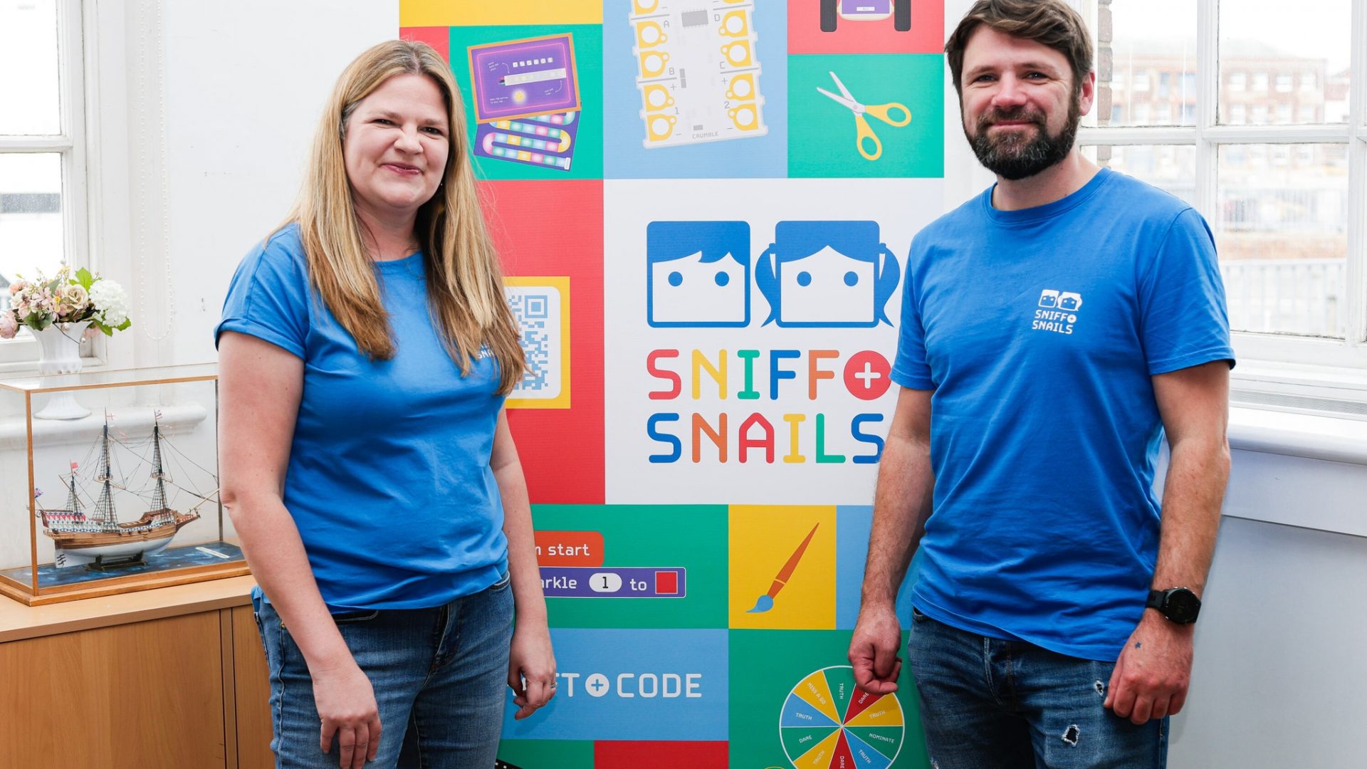 Two people in blue Sniff & Snails shirts stand smiling in front of a colorful Sniff & Snails banner with educational icons in a bright room with windows and a model ship on a cabinet.