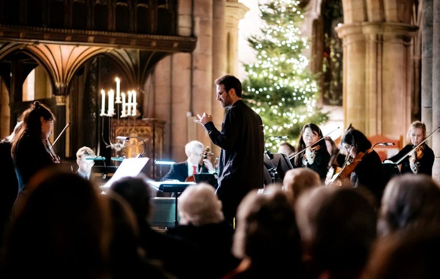 A conductor leads an orchestra performing in a warmly lit church decorated with a glowing Christmas tree, as an audience watches attentively.