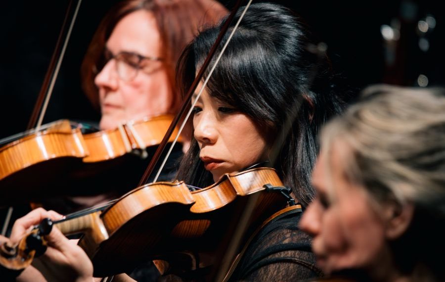 Three women play violins in an orchestra, focused and concentrated, with blurred background musicians.