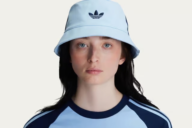 A person with long dark hair wears a light blue Adidas bucket hat and a matching light blue Adidas shirt with navy stripes, looking straight at the camera against a plain background.