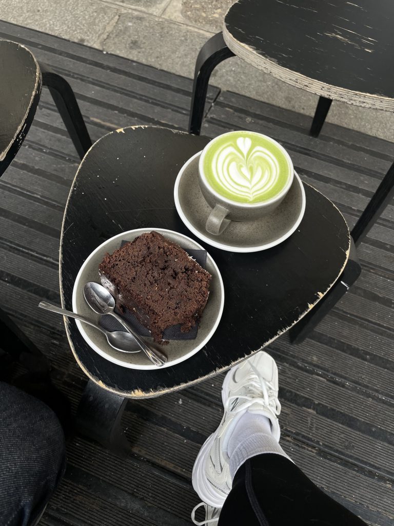A slice of chocolate cake with two spoons in a bowl and a cup of matcha latte with a heart leaf latte art, on black tables outside. A person’s crossed legs in white sneakers and socks are visible below.