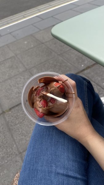 A person sitting outdoors in jeans holds a cup of chocolate ice cream topped with strawberries and a white plastic spoon. A sidewalk and part of a green table are visible in the background.