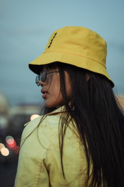 A person with long dark hair, wearing a yellow bucket hat, glasses, and a yellow jacket, looks over their shoulder outdoors at dusk with blurred city lights in the background.