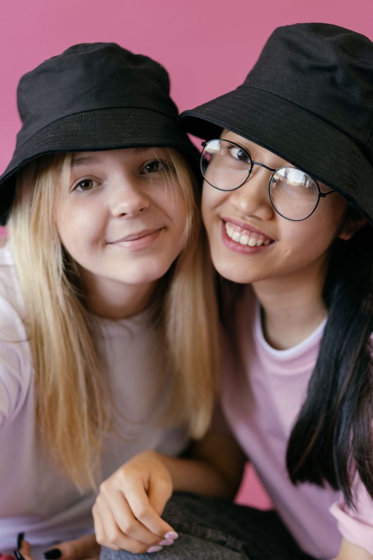 Two young women smiling together in front of a pink background. Both are wearing black bucket hats; one has blonde hair and the other has dark hair and glasses. They appear to be taking a selfie and look happy.