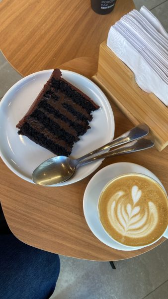 A slice of chocolate layer cake on a white plate with a fork and spoon, next to a cup of latte with latte art, placed on a wooden table beside a napkin holder.