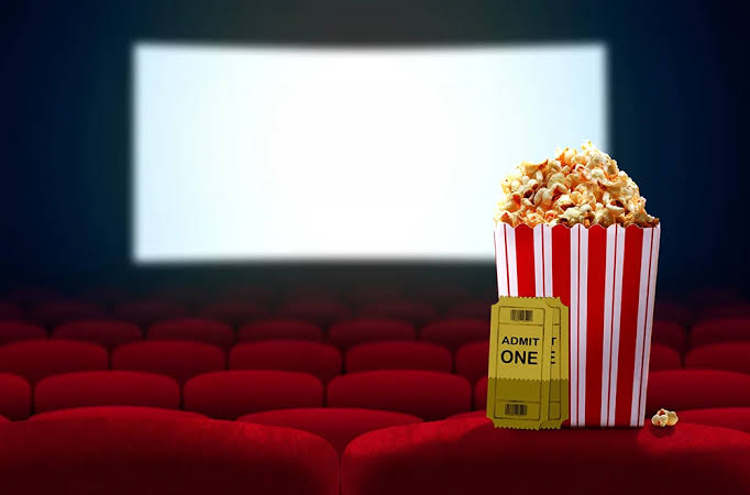 A striped popcorn container and a movie ticket sit on red theater seats in front of a blank, illuminated cinema screen.