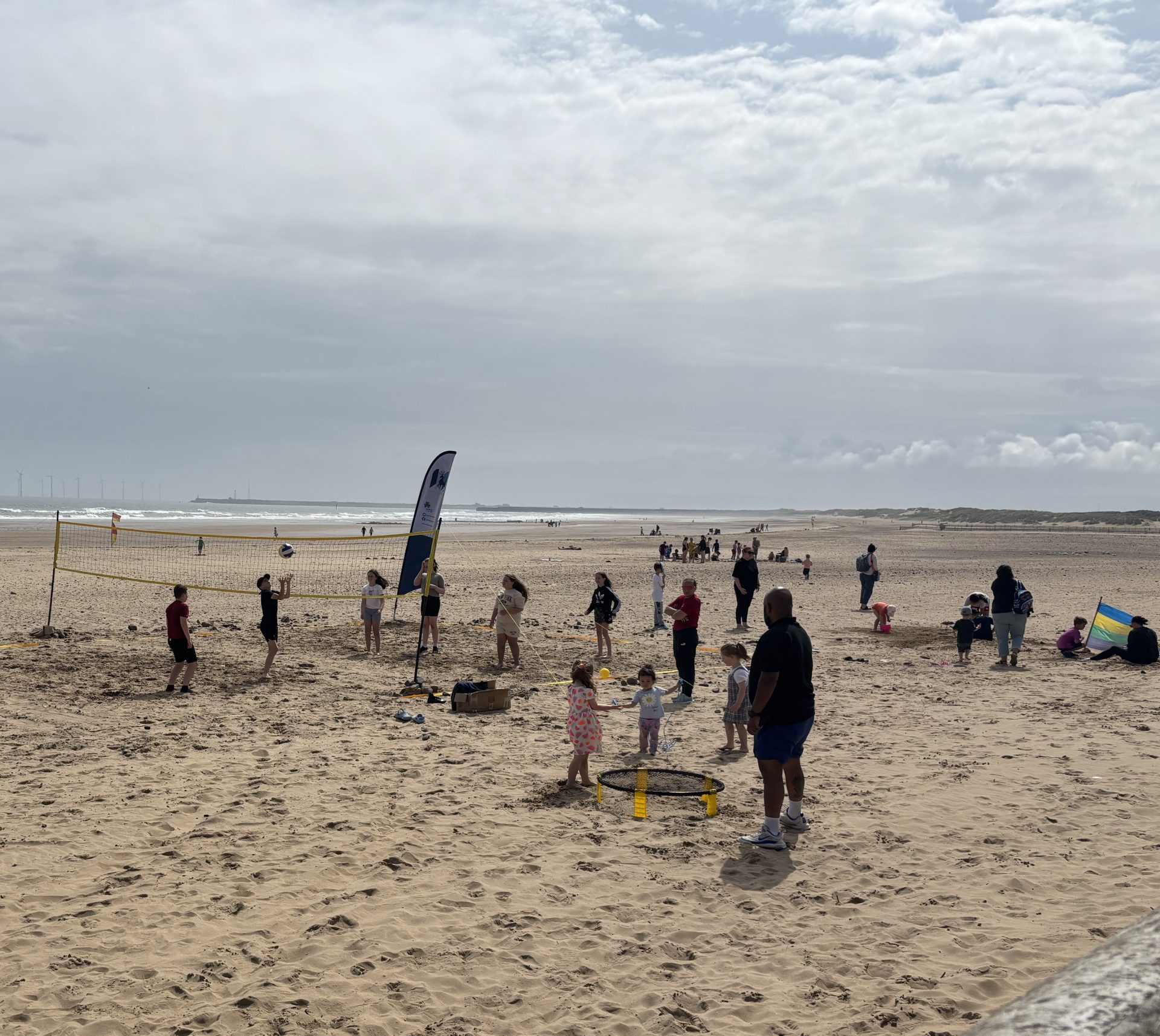 People, including children, play games on a sandy beach under a partly cloudy sky. A volleyball net and a flag are set up, with the sea in the background and more people scattered along the shore.