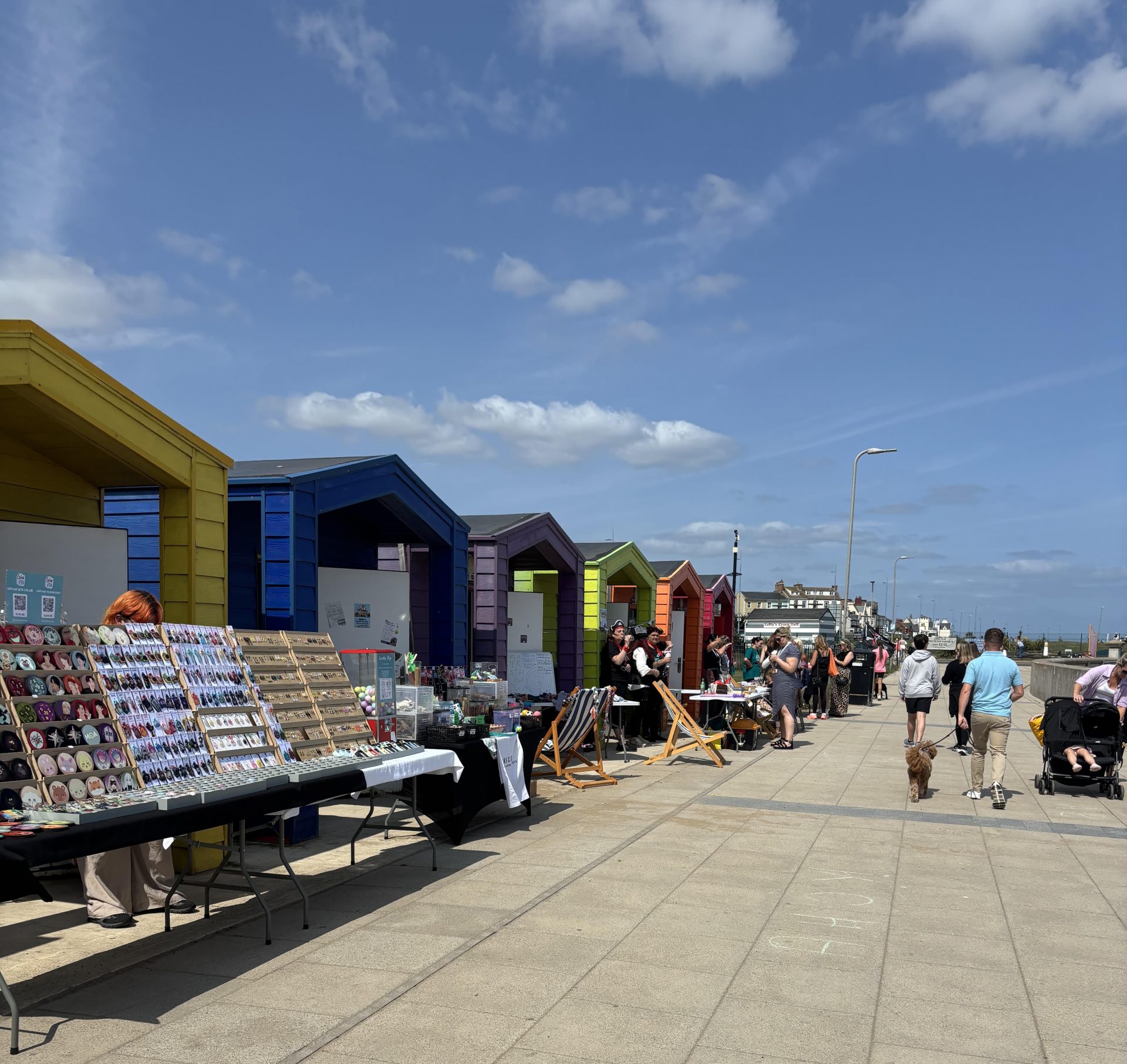 Outdoor market with stalls in front of colorful beach huts; people browse the goods while others walk along the paved path under a partly cloudy sky.