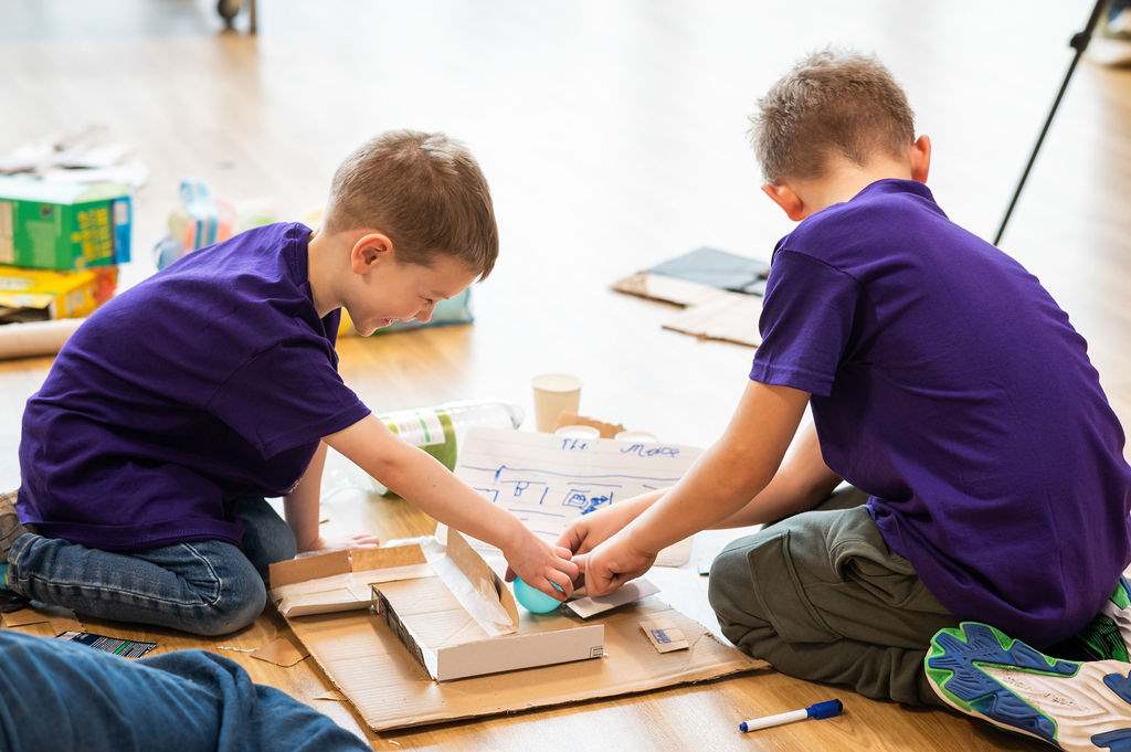 Two young boys in purple shirts sit on the floor, smiling and playing with cardboard and craft materials, creating a project together. Other scattered supplies are also visible around them.