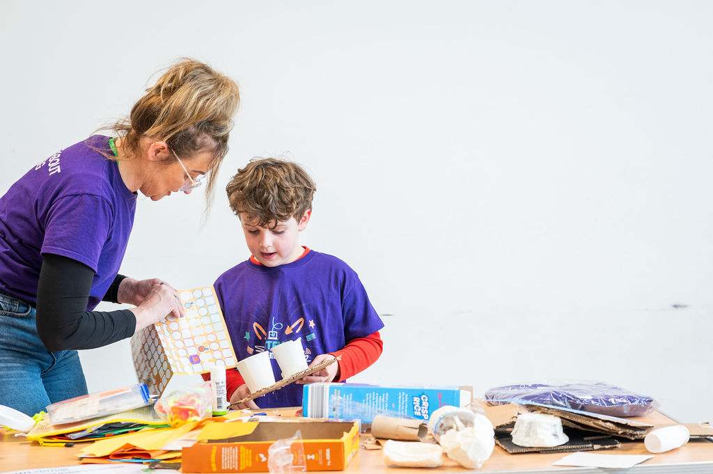 An adult and a child, both wearing purple shirts, work together on a craft project with various recycled materials on a table in front of them.