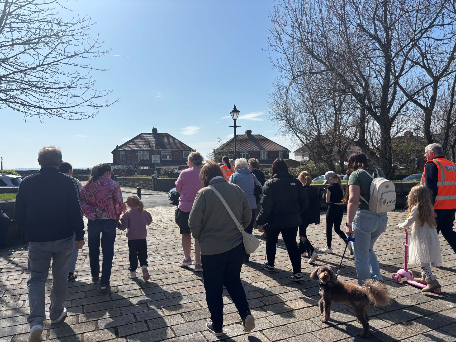 A group of people, including children and a dog, walk together on a sunny day along a paved path, with houses, trees, and a lamppost visible in the background.