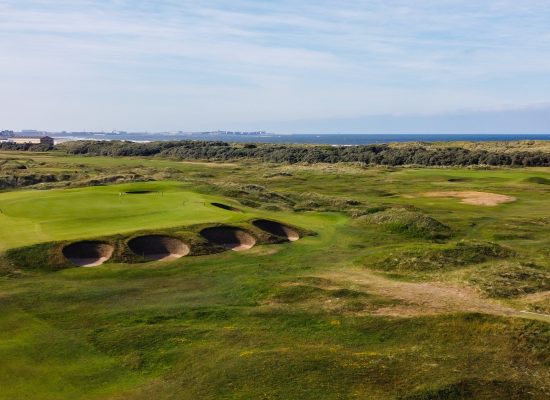 A scenic view of a coastal golf course with several sand bunkers, rolling green fairways, grassy mounds, and the sea visible in the background under a partly cloudy sky.