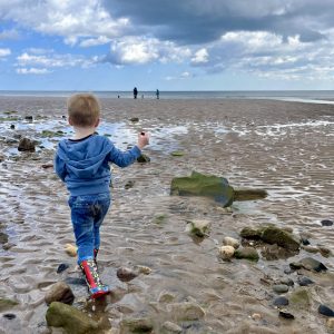 A young child wearing a blue hoodie and colourful boots walks on a rocky, sandy beach toward the sea under a cloudy sky. There are a few people in the distance near the water.