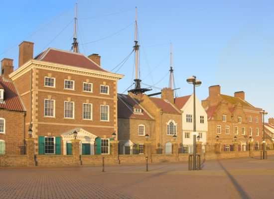 A row of historic brick buildings with gabled roofs and green shutters line a spacious, sunlit plaza; a tall ship’s masts rise behind the central building under a clear blue sky.