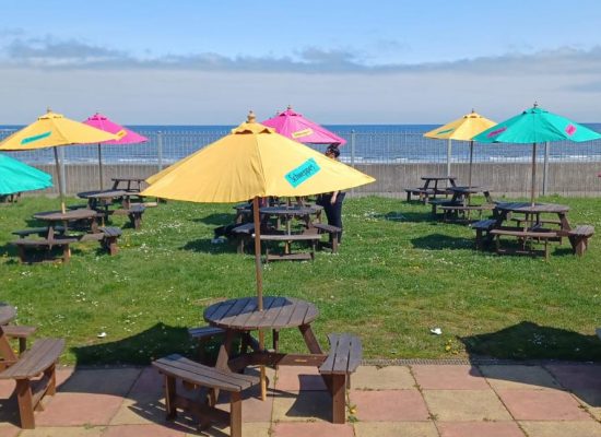 Several wooden picnic tables with colourful umbrellas are arranged on a grassy area near a seaside, with a clear view of the sea and a partly cloudy sky in the background. Two people sit at a table in the foreground.
