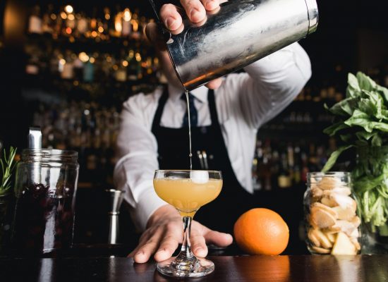 Man wearing shirt, tie and apron pours a cocktail from a metal shaker.