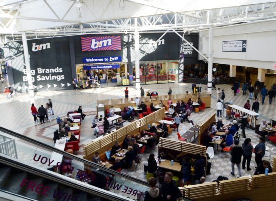A busy shopping mall food court with people seated at tables and walking around; in the background, a large B&M store entrance displays a colourful Welcome to B&M sign. Bright natural light fills the space.